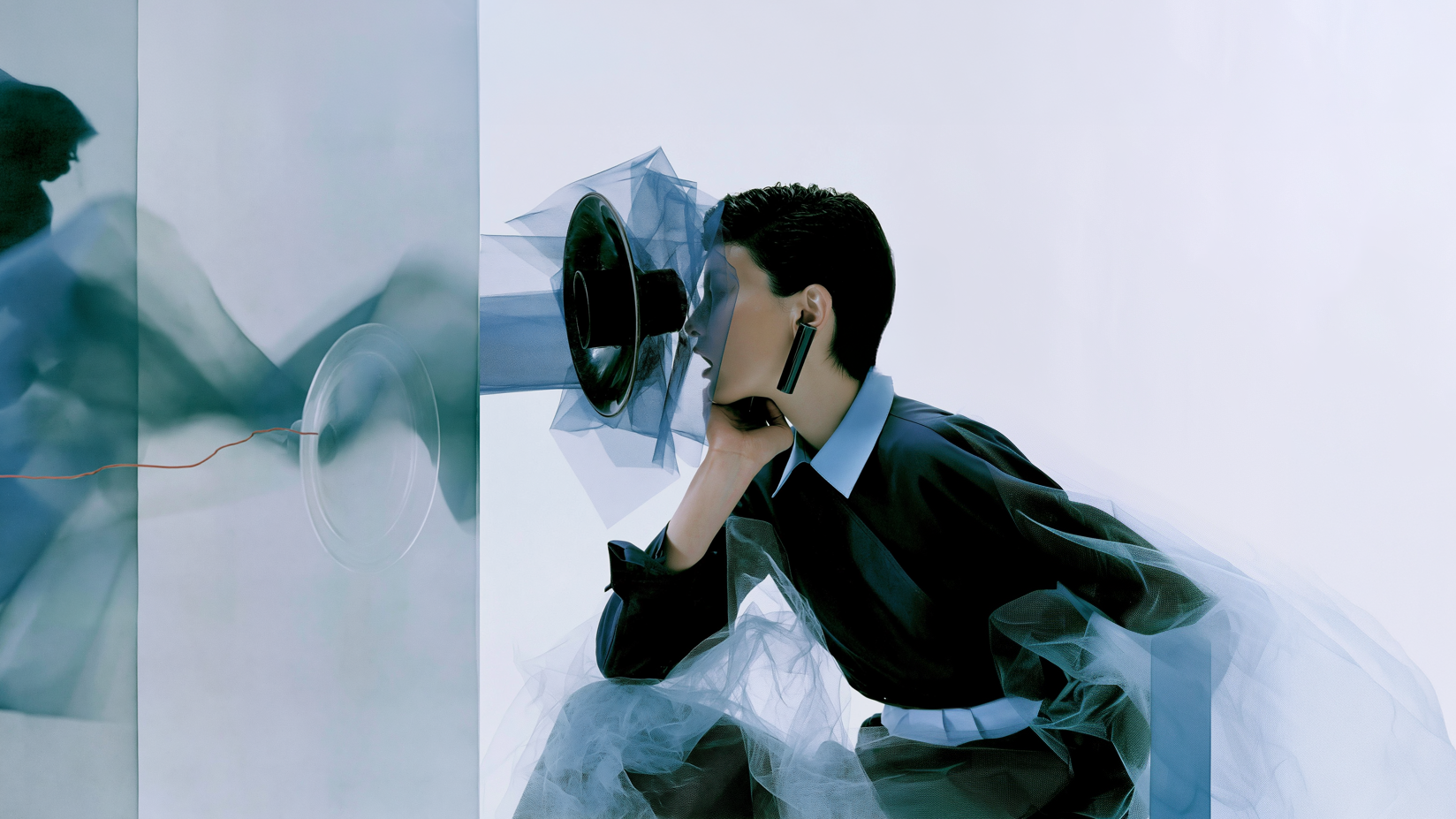 A woman listening to a sculptural megaphone amid translucent blue fabric.