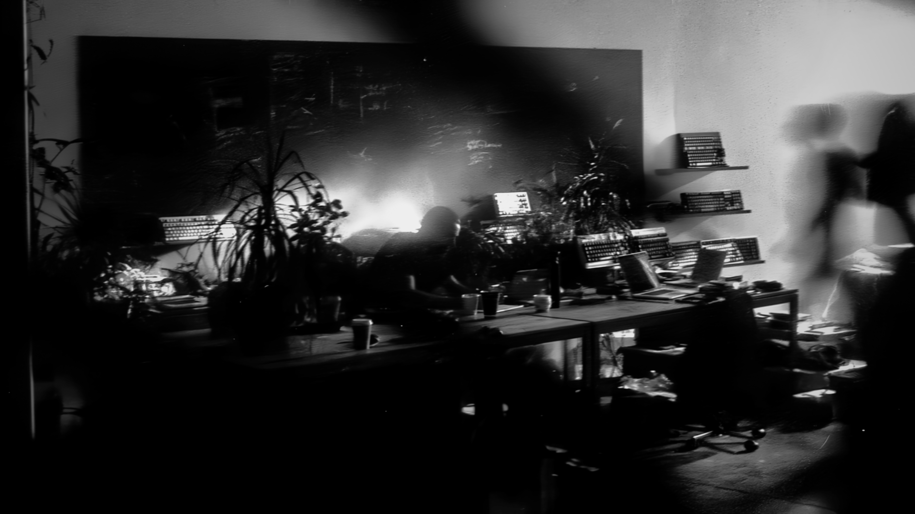 A motion-softened black-and-white creative studio filled with desks, plants, keyboards, laptops, and anonymous creators.